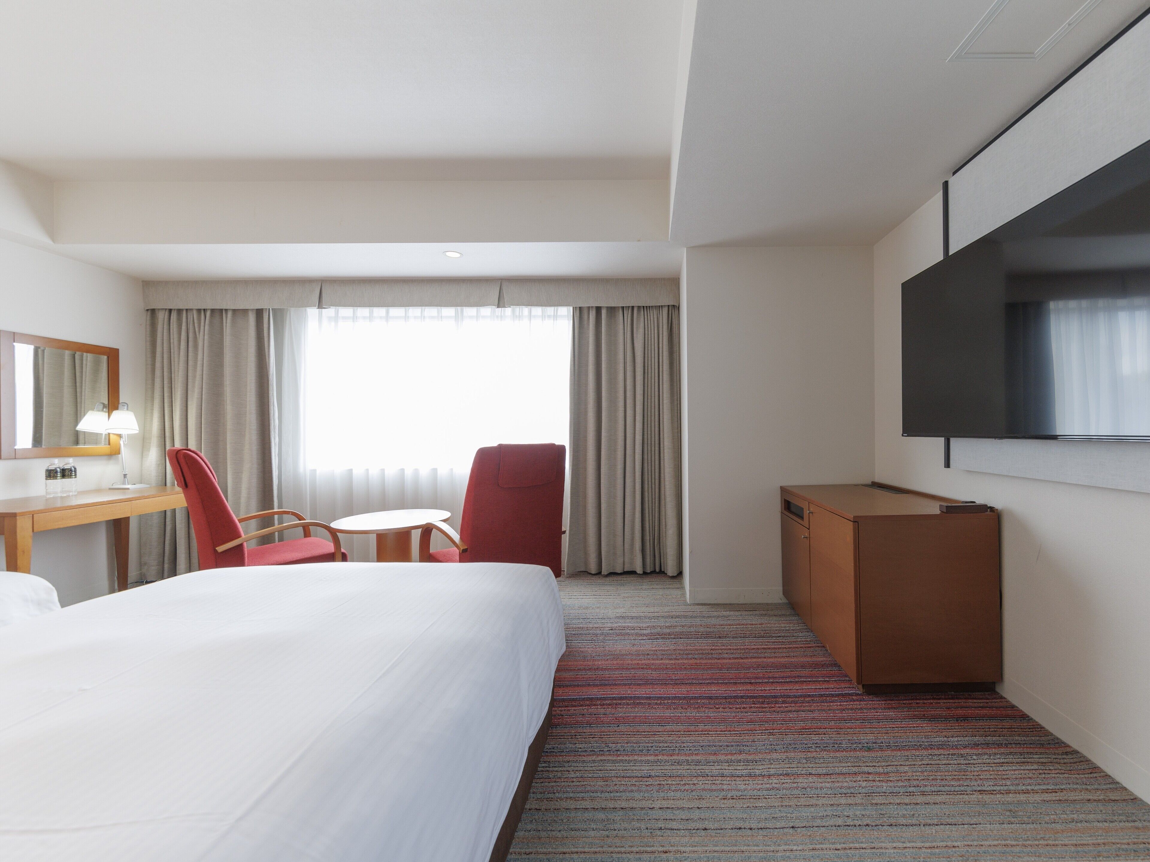Habitación de hotel con cama, escritorio con lámpara, televisor, una silla roja y ventana con cortinas.
