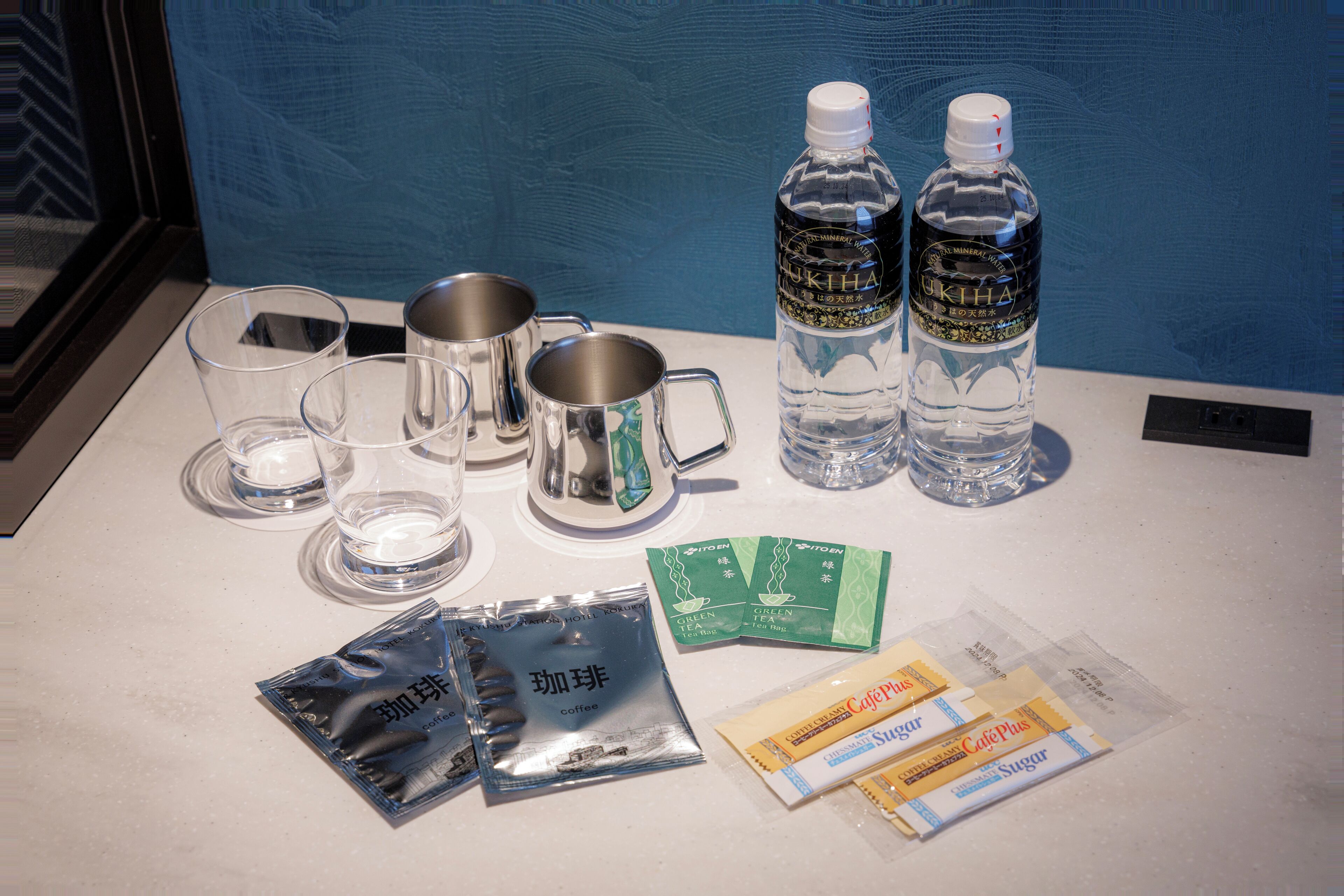 Una mesa con dos botellas de agua, dos vasos transparentes, tres tazas de acero inoxidable y sobres de café y azúcar.