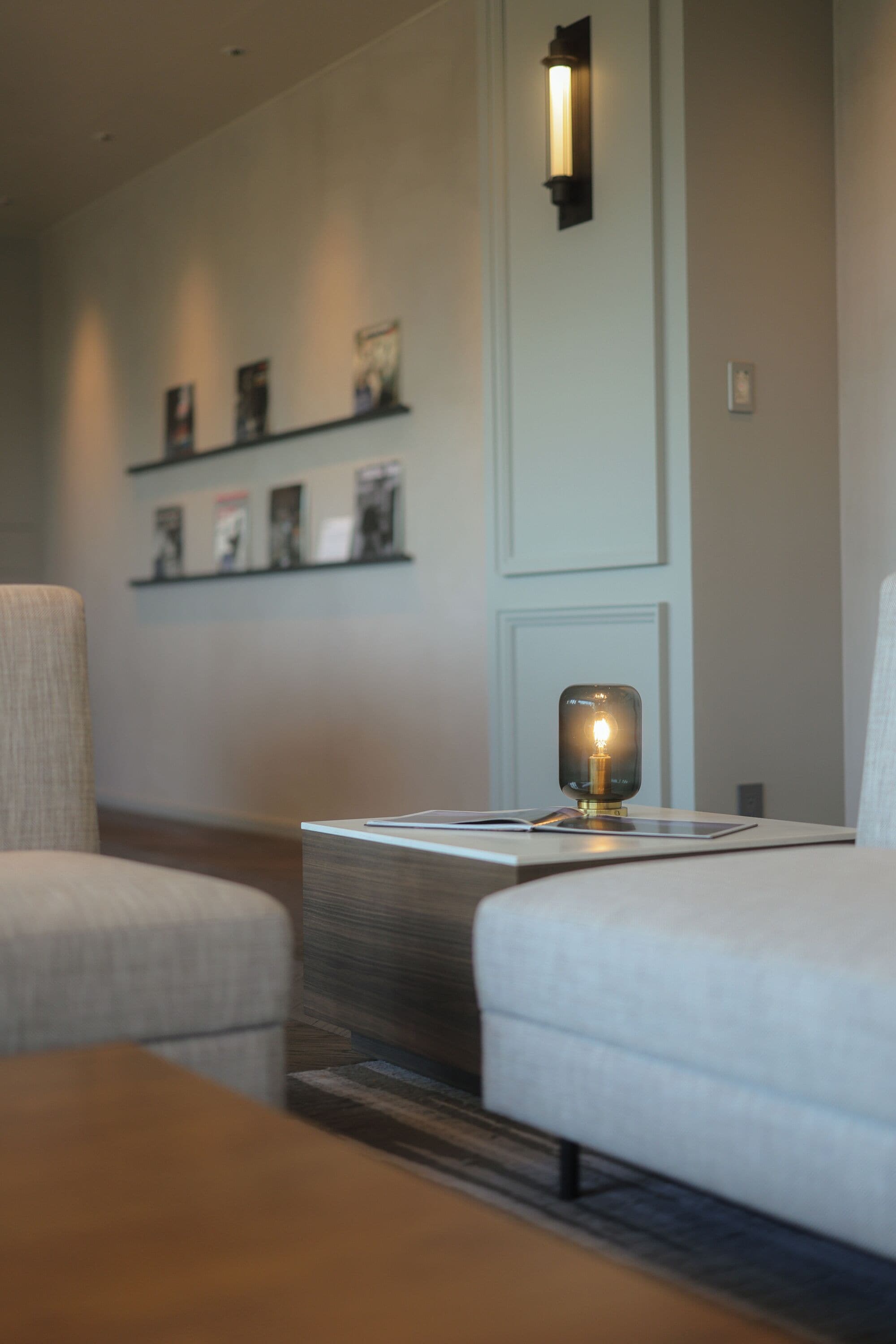 A modern hotel room with a sofa, a side table with a lamp, and a wall-mounted shelf with framed pictures.