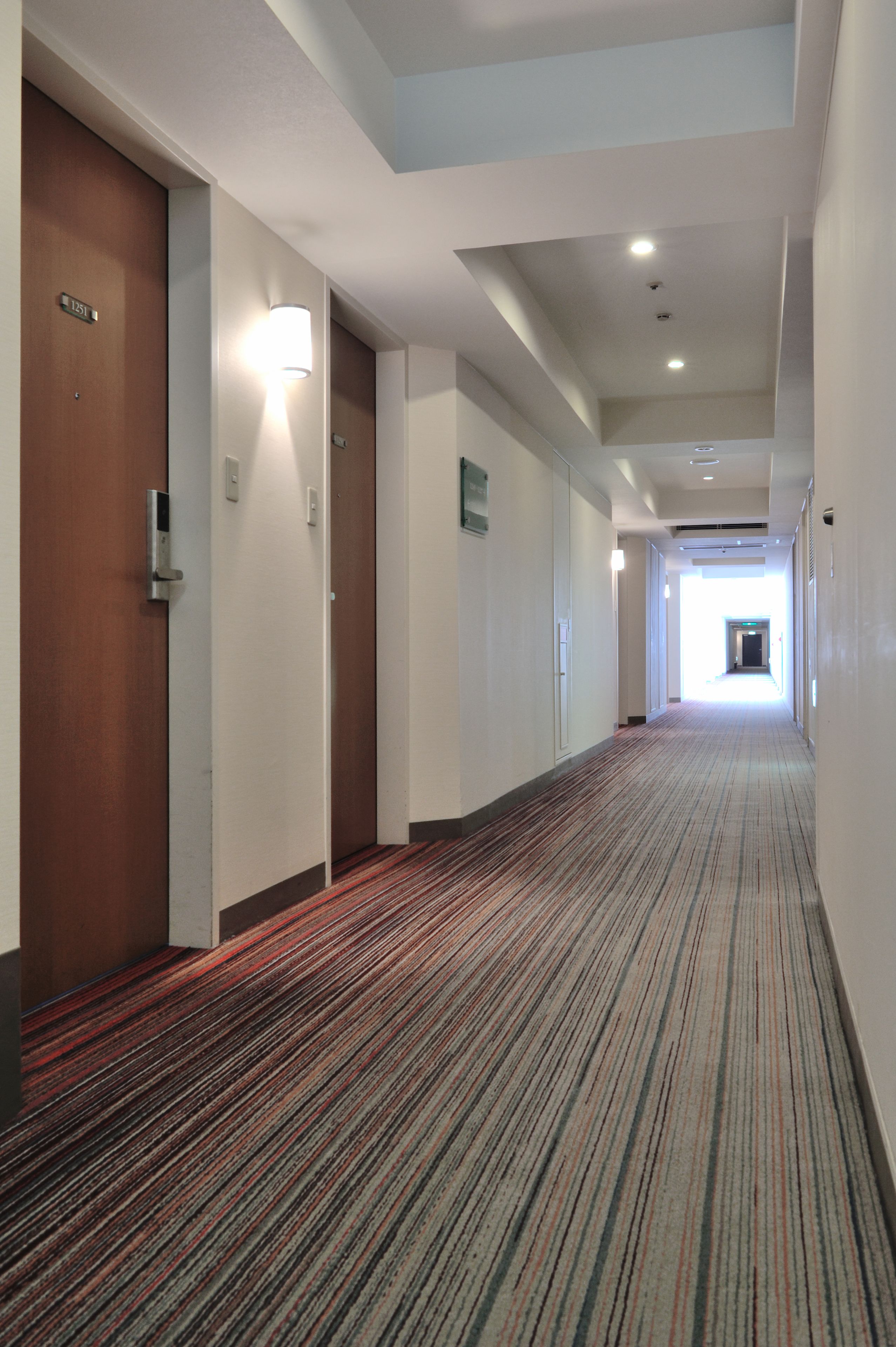 Un couloir d'hôtel avec des revêtements de sol en moquette, des portes en bois et un éclairage encastré.