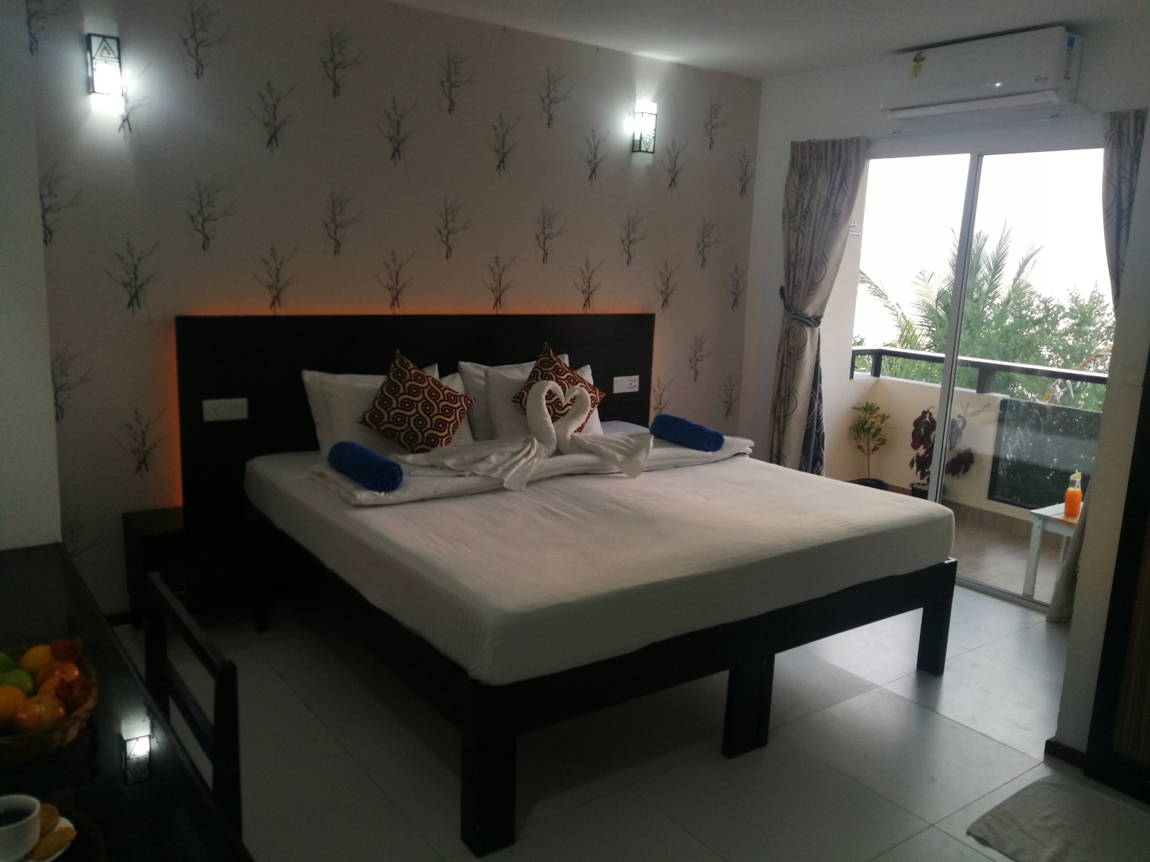 A neatly made bed with patterned pillows, a bedside table with a bottle, and a view of the outdoors through a balcony.