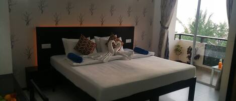 A neatly made bed with patterned pillows, a bedside table with a bottle, and a view of the outdoors through a balcony.