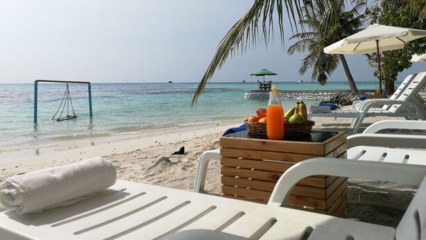 Un espacio de relax frente al mar con sillas blancas, una mesa de madera con frutas y una botella de jugo de naranja, y una vista despejada del océano.
