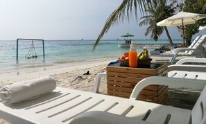 Un salon de plage avec des chaises longues blanches, une table en bois sur laquelle sont disposés des fruits et une bouteille de jus d’orange, et une vue dégagée sur l’océan.
