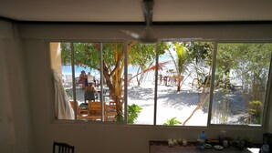 Una habitación con un amplio ventanal con vista a una playa, mobiliario exterior y palmeras.