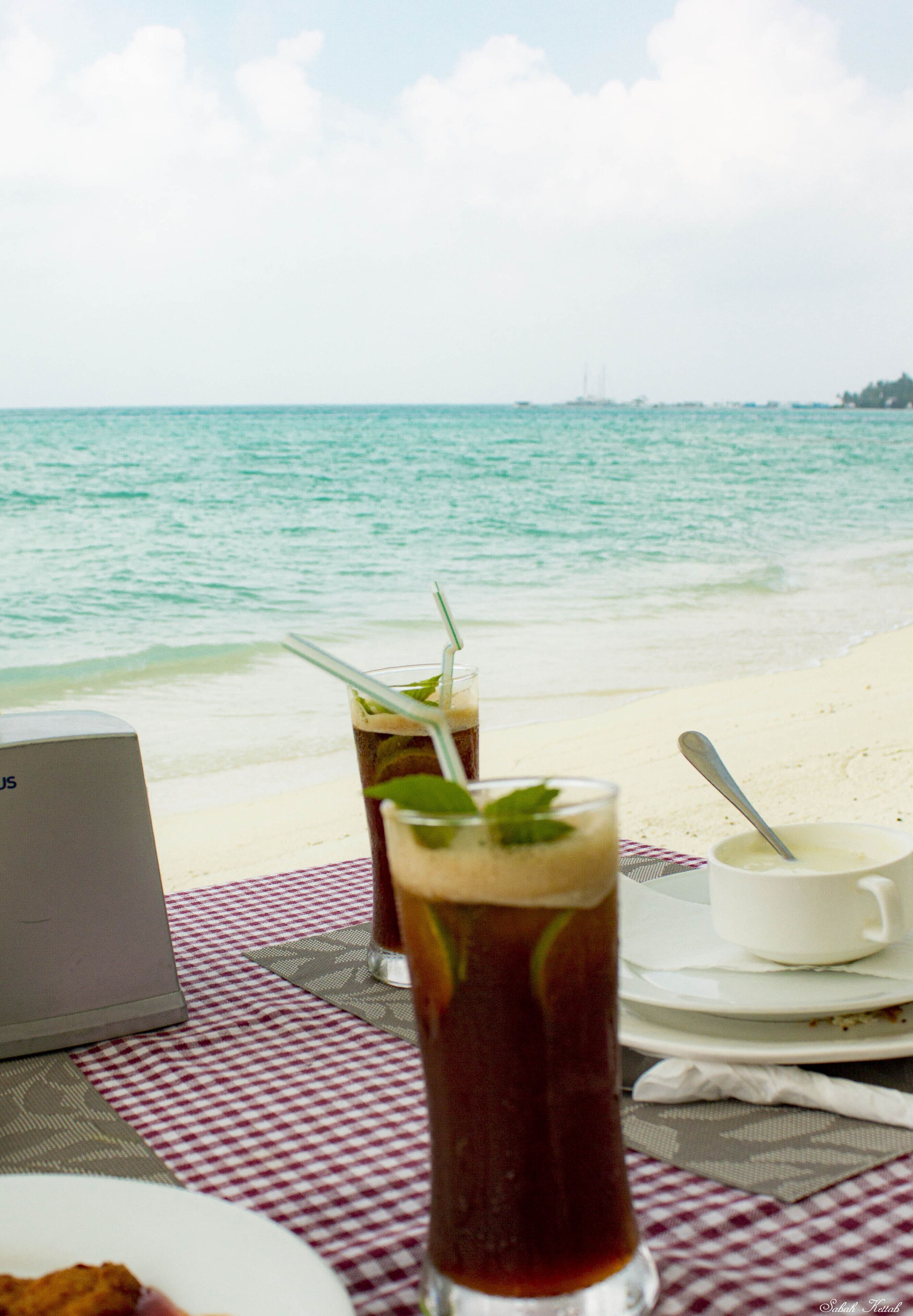 A beachside table with drinks, a laptop, and a white cup on a checkered tablecloth.