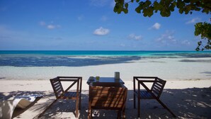 Vista de una playa con dos sillas de madera y una mesa, cielo azul despejado y mar turquesa.