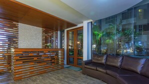 A modern reception area with a wooden reception desk, a leather sofa, and a large window with a city view. - Express Inn (Hulhumalé)