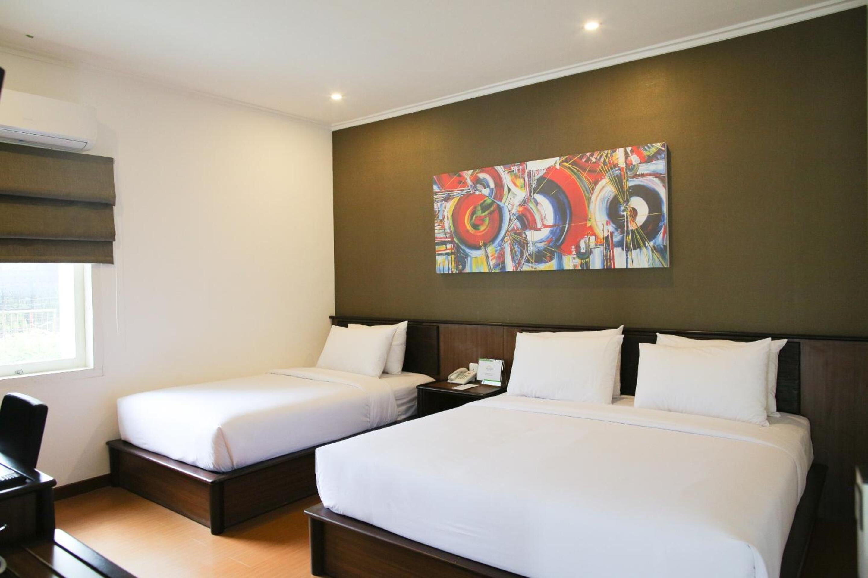 A hotel room with two single beds, a large abstract painting on the wall, and a window with blinds.