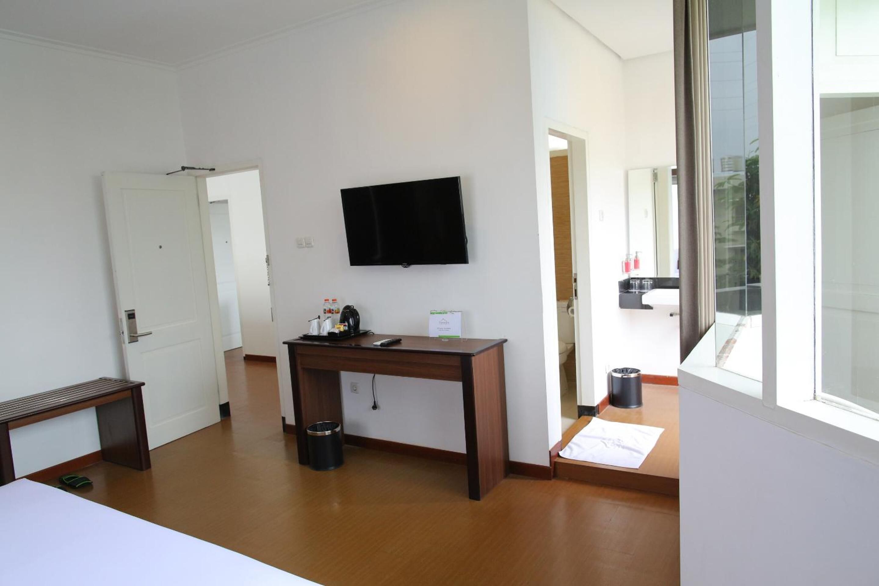 A hotel room with a wooden desk, a flat-screen TV mounted on the wall, a small trash bin, and a window with a view of the outside.