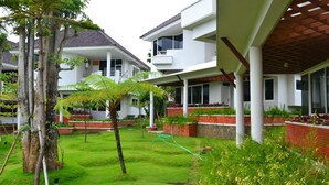A modern building with a covered outdoor area, a well-maintained lawn, and a variety of plants. - Samara Resort (Batu)