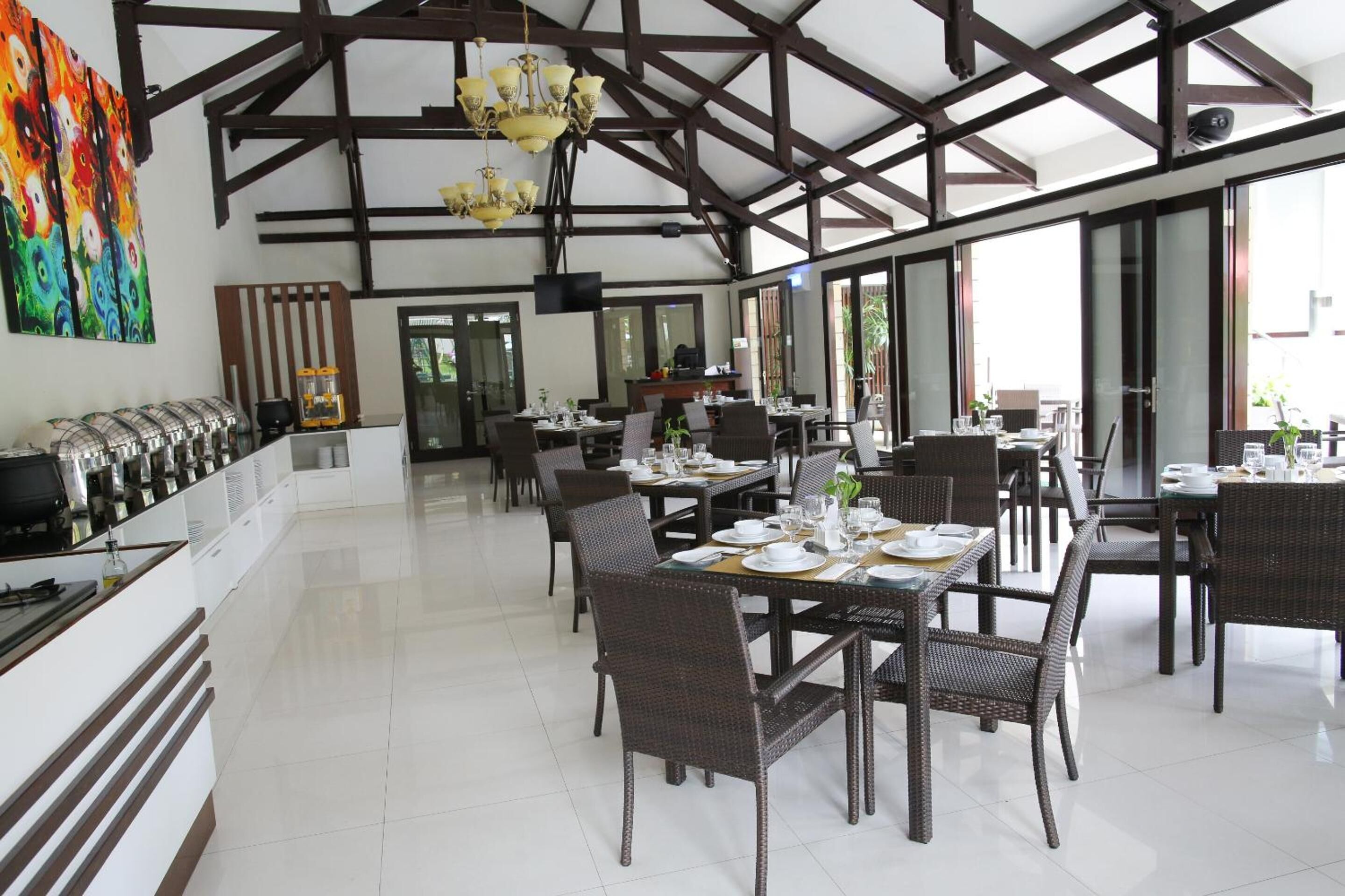 Uma ampla sala de jantar com pé direito alto e vigas aparentes, com várias mesas cobertas por toalhas brancas e cadeiras de vime escuro.