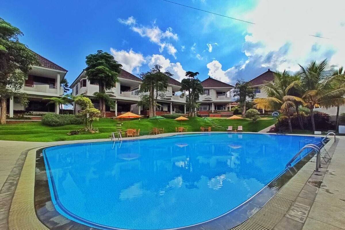 A swimming pool surrounded by buildings with balconies, a well-maintained lawn, and palm trees.