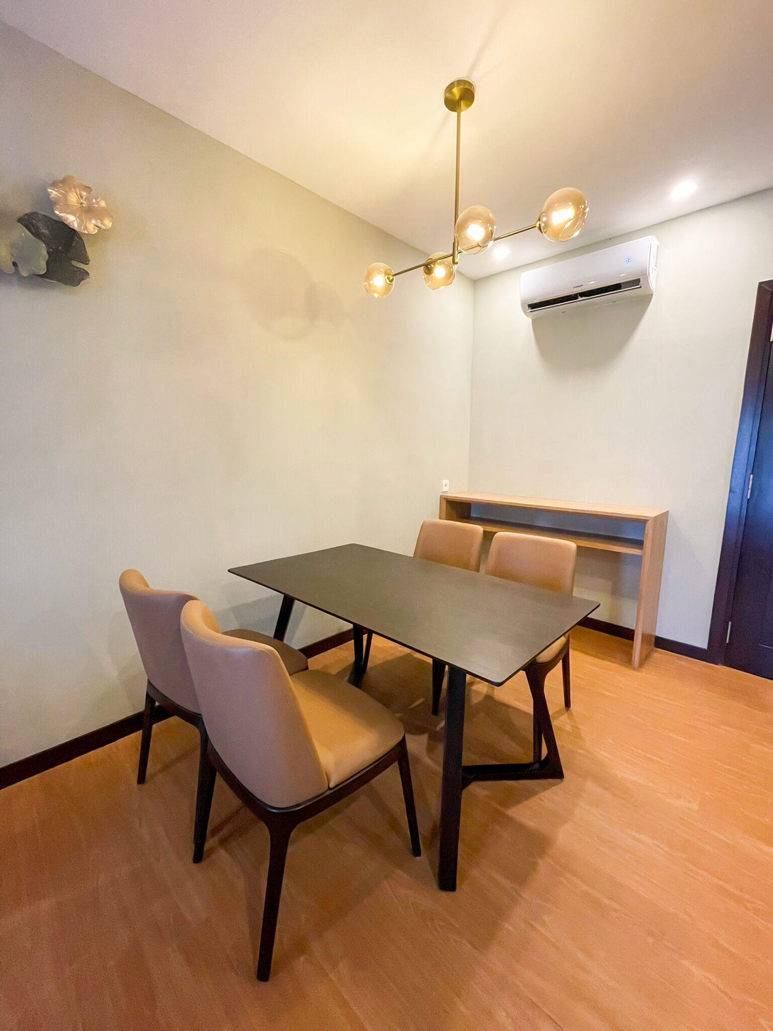 Una habitación con una mesa de comedor rectangular, cuatro sillas, un aire acondicionado de pared y una lámpara en el techo.