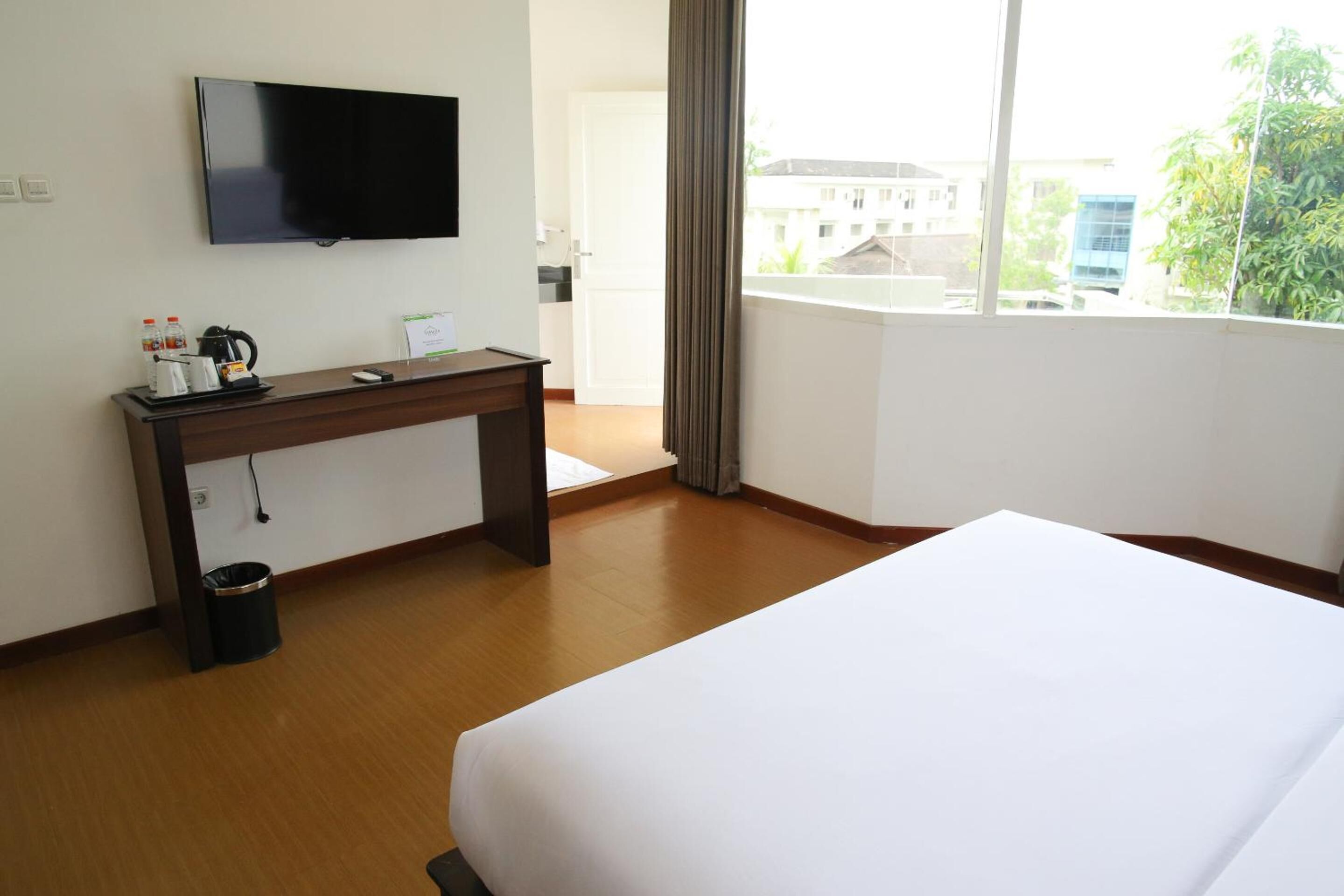 Habitación de hotel con una cama, un escritorio con televisor, un cesto de basura y ventana con vistas a edificios y árboles.