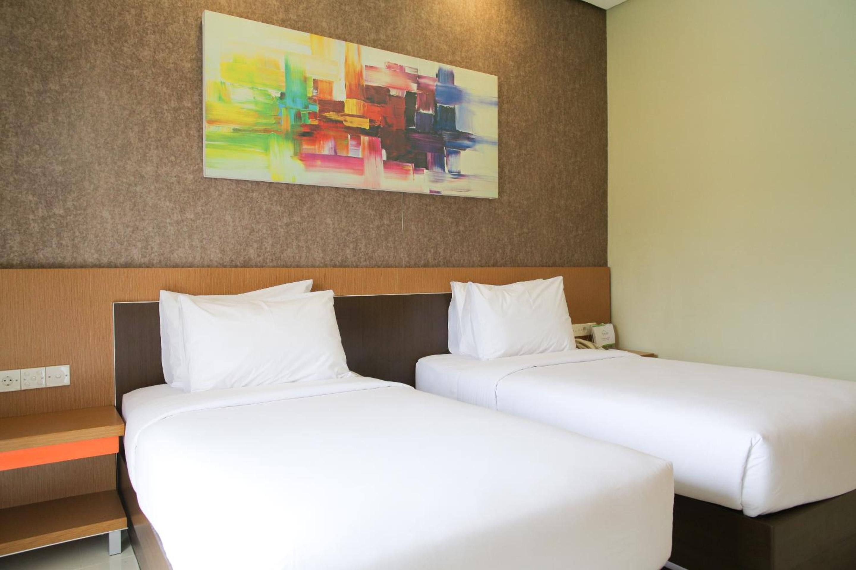 A hotel room with two single beds, a wooden headboard, a nightstand, and a colorful abstract painting on the wall.