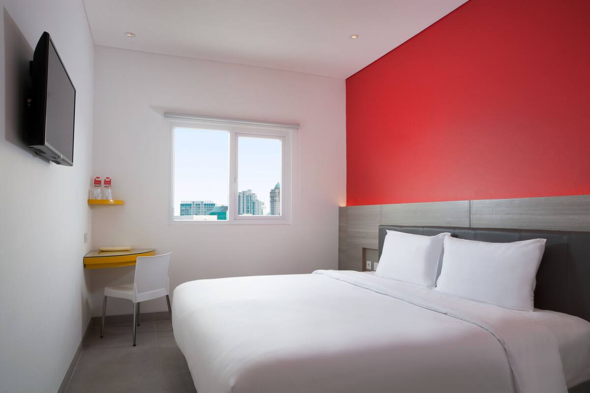 Habitación de hotel con cama, escritorio, silla, televisor montado en la pared y ventana con vista a la ciudad.