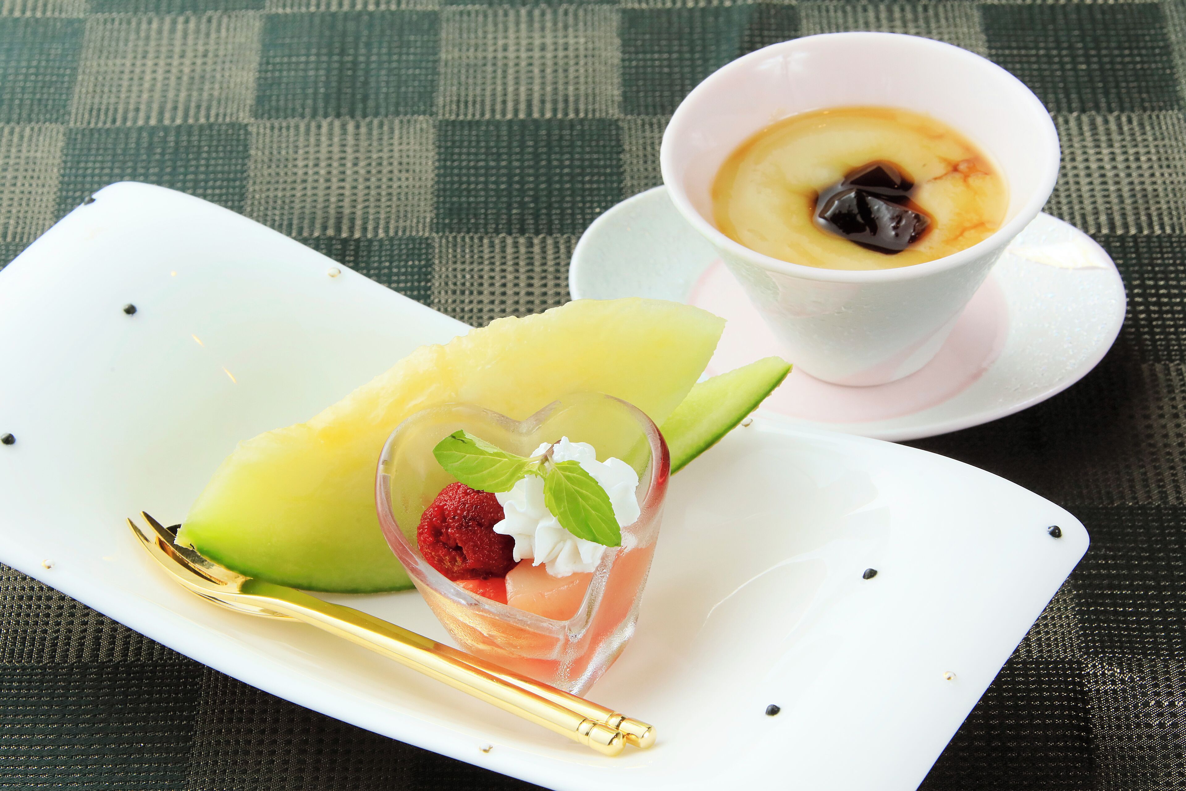 A dessert plate with a heart-shaped dessert, a slice of mango, and a raspberry, accompanied by a cup of coffee on a checkered tablecloth.