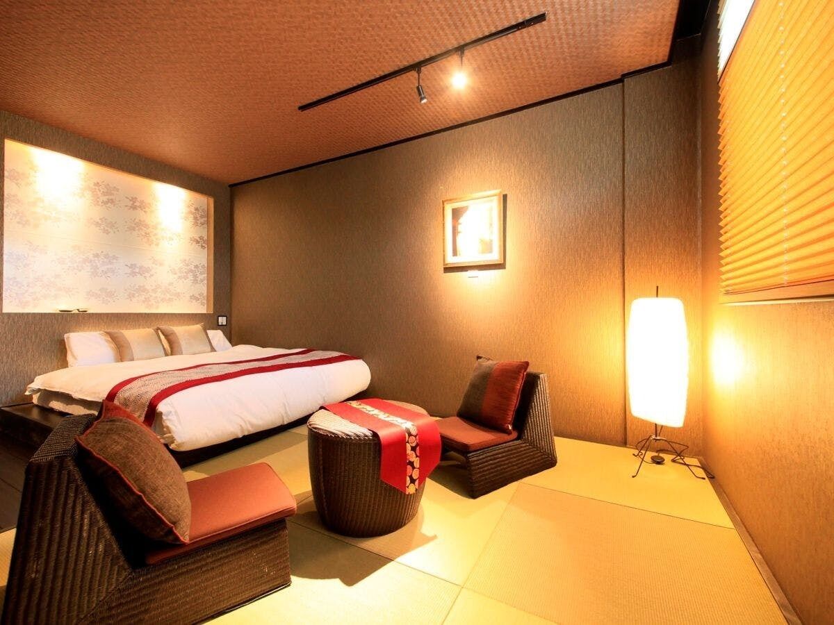 A hotel room with a bed, two chairs, a small ottoman, a wall-mounted light, and a framed picture.