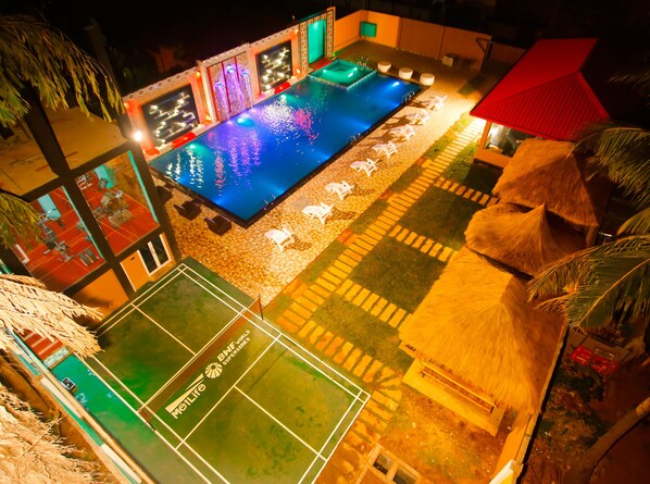 Una piscina con suelo a cuadros, una cancha de tenis y una estructura cubierta de paja.
