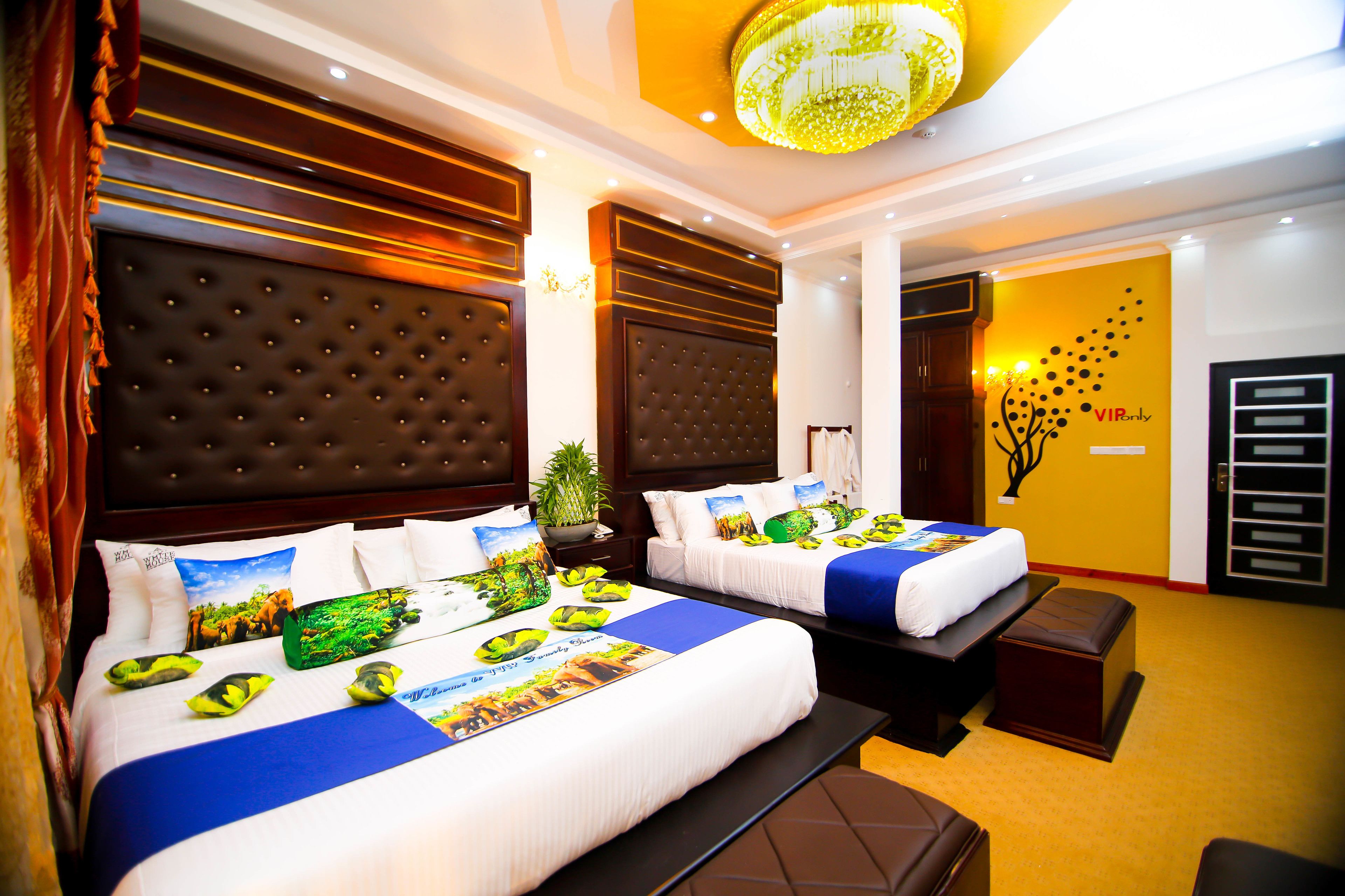A hotel room with two beds, each with a blue and white bedspread, a large tufted headboard, and a chandelier above.