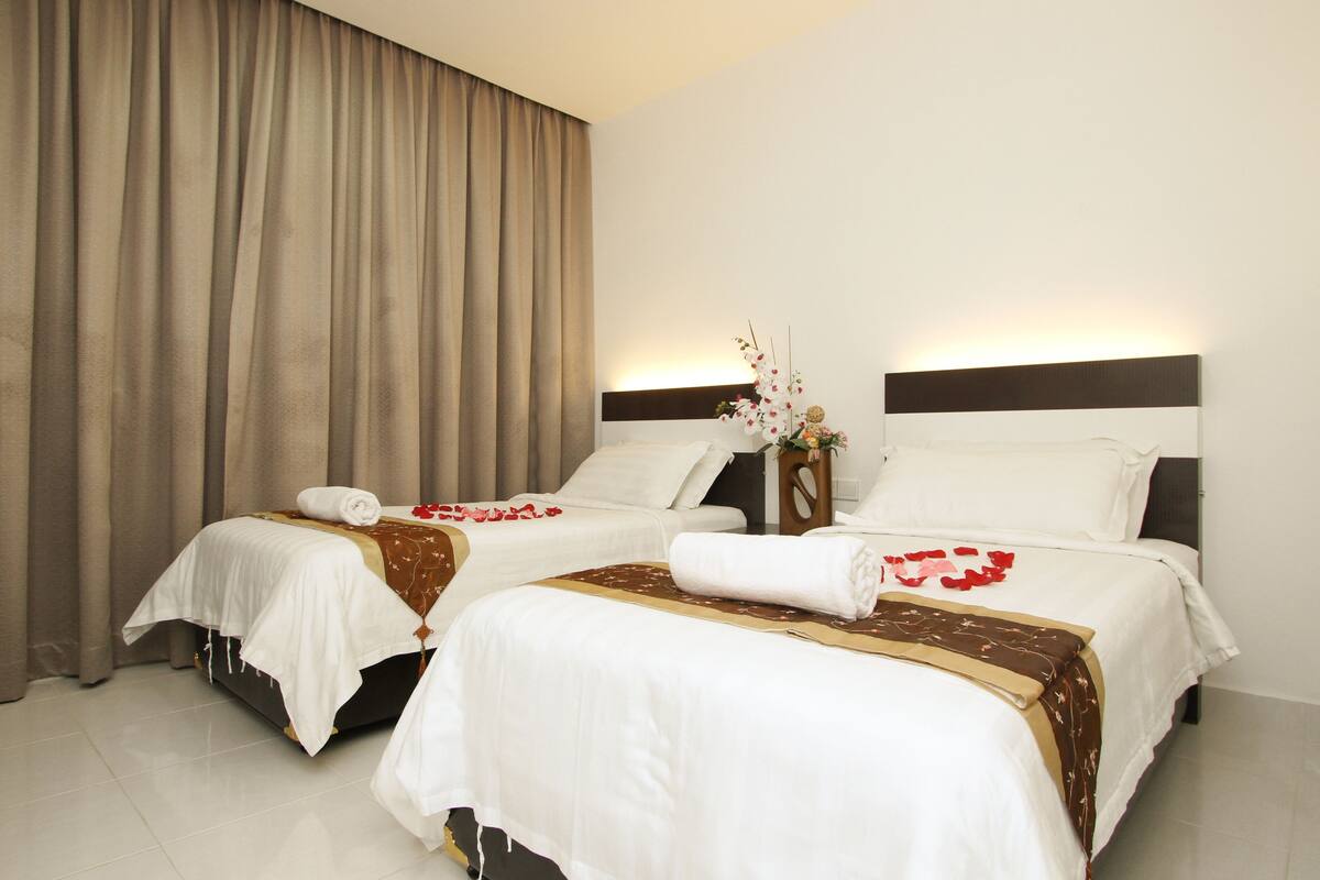 Quarto de hotel com duas camas, cortinas bege e um vaso com flores.