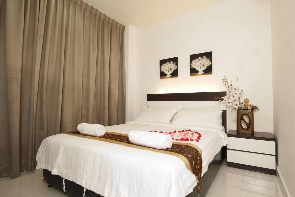 Cama bem arrumada com lençóis brancos, um corredor decorativo marrom e duas toalhas enroladas. Há duas pinturas de flores penduradas na parede, uma mesa de cabeceira com um vaso de flores e uma janela grande com cortinas.