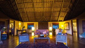 A spacious room with a high thatched ceiling, a large cushioned sofa, two wicker chairs, a coffee table, and a dining table with chairs. - Elewana Tortilis Camp (Amboseli)