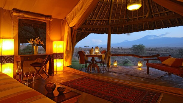 A cozy room with a bed, a dining table, and chairs. There is a view of a mountain and a desert landscape. - Elewana Tortilis Camp (Amboseli)