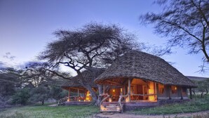 A thatched-roof lodge with a stone pathway leading to the entrance, surrounded by natural vegetation. - Elewana Tortilis Camp (Amboseli)