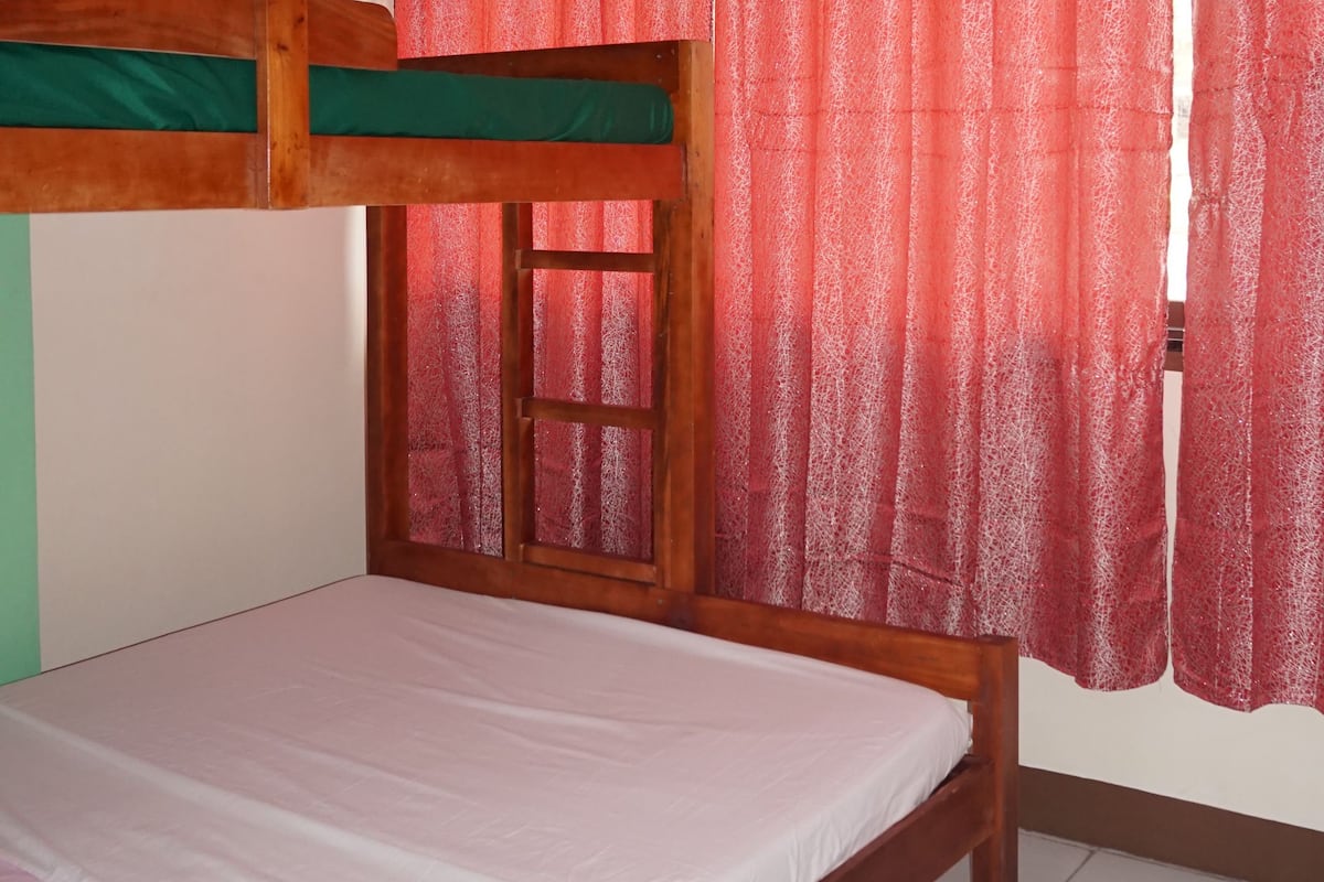 A single-bed room with a wooden headboard, a window with red curtains, and a green wall.