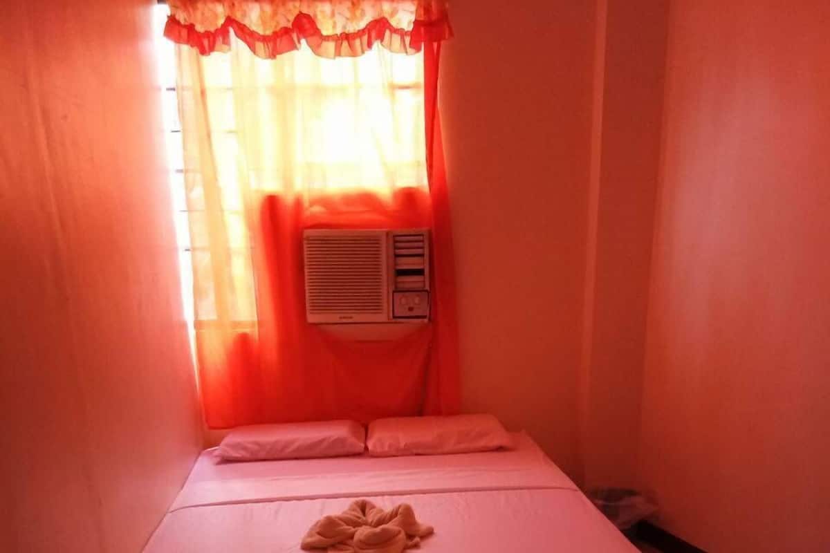 A small, single-bed room with a mattress, pillows, and a blanket. There is a window with sheer curtains and a wall-mounted air conditioner.