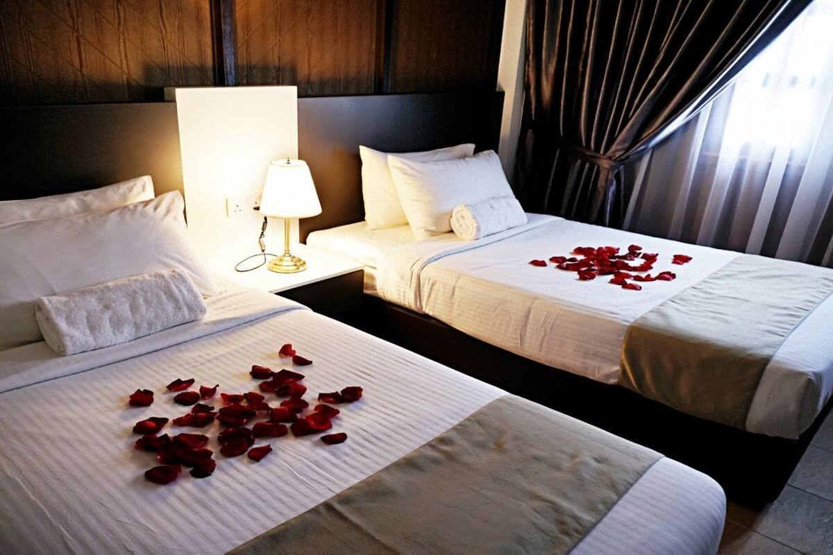 Two beds with white linens and red rose petals, a bedside lamp, and a dark wooden headboard.