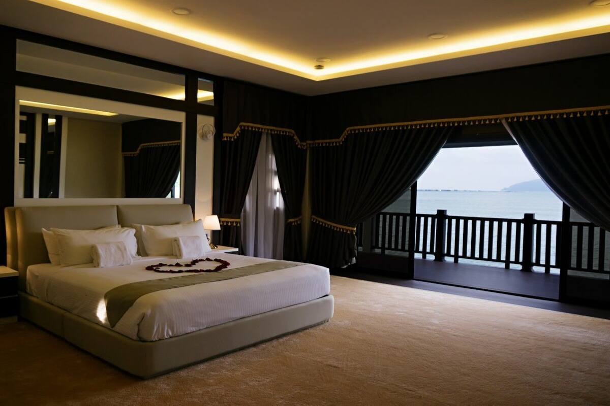 A large bed with a headboard, a bedside table with a lamp, and a view of the sea through a balcony with curtains.