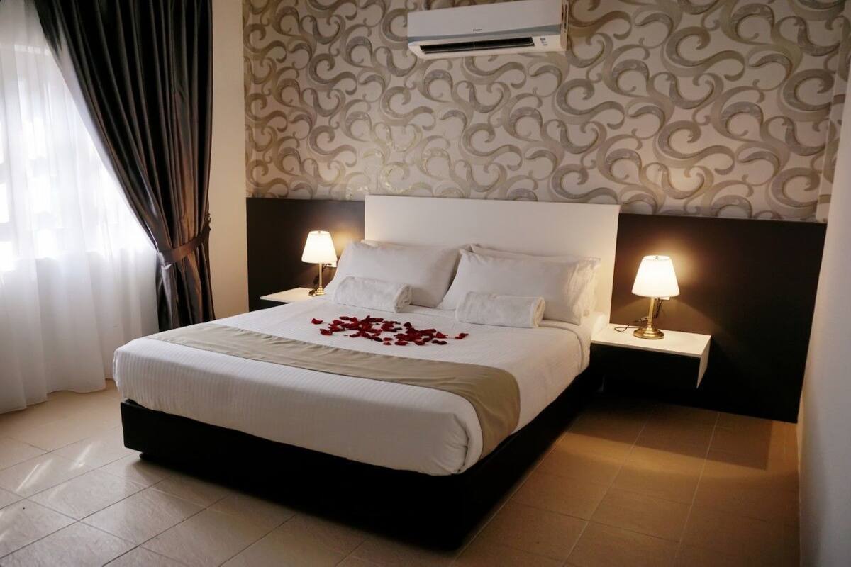 A hotel room with a large bed, two bedside tables with lamps, a patterned wallpaper, and an air conditioning unit on the wall.