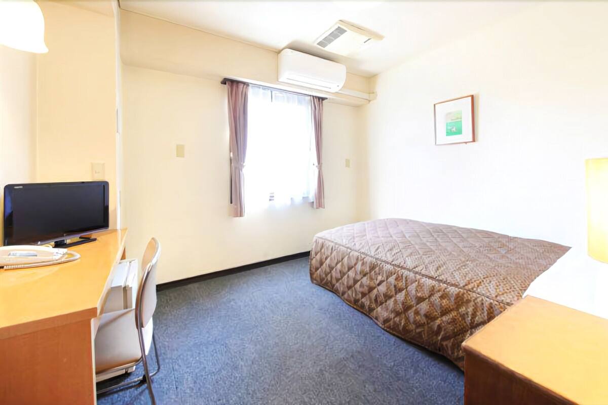 Habitación de hotel con cama, escritorio con computadora, silla, equipo de aire acondicionado y ventana con cortinas.