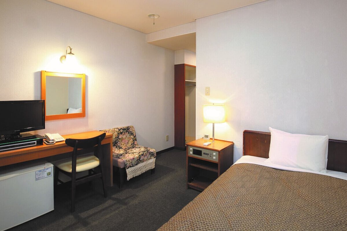 Habitación de hotel con cama, escritorio con televisor, silla, lámpara de escritorio y refrigerador pequeño.