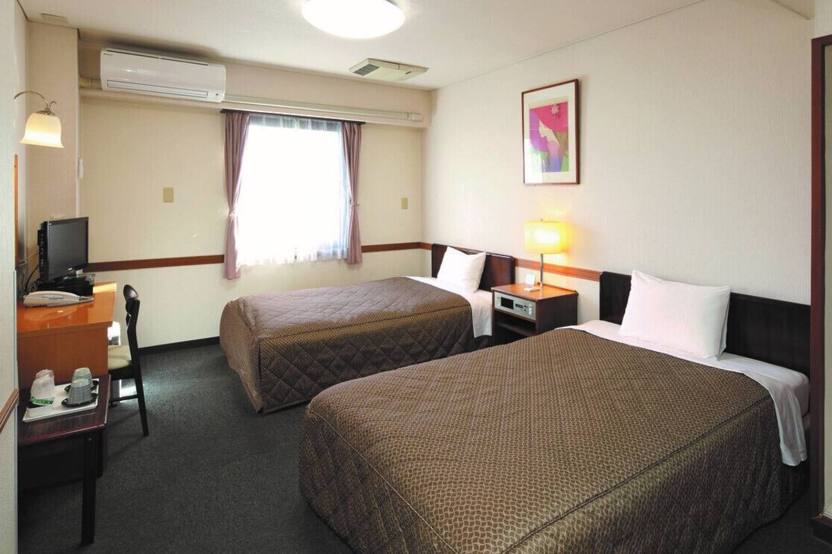 Habitación de hotel con dos camas, un escritorio con computadora, una silla, una lámpara y una ventana con cortinas.