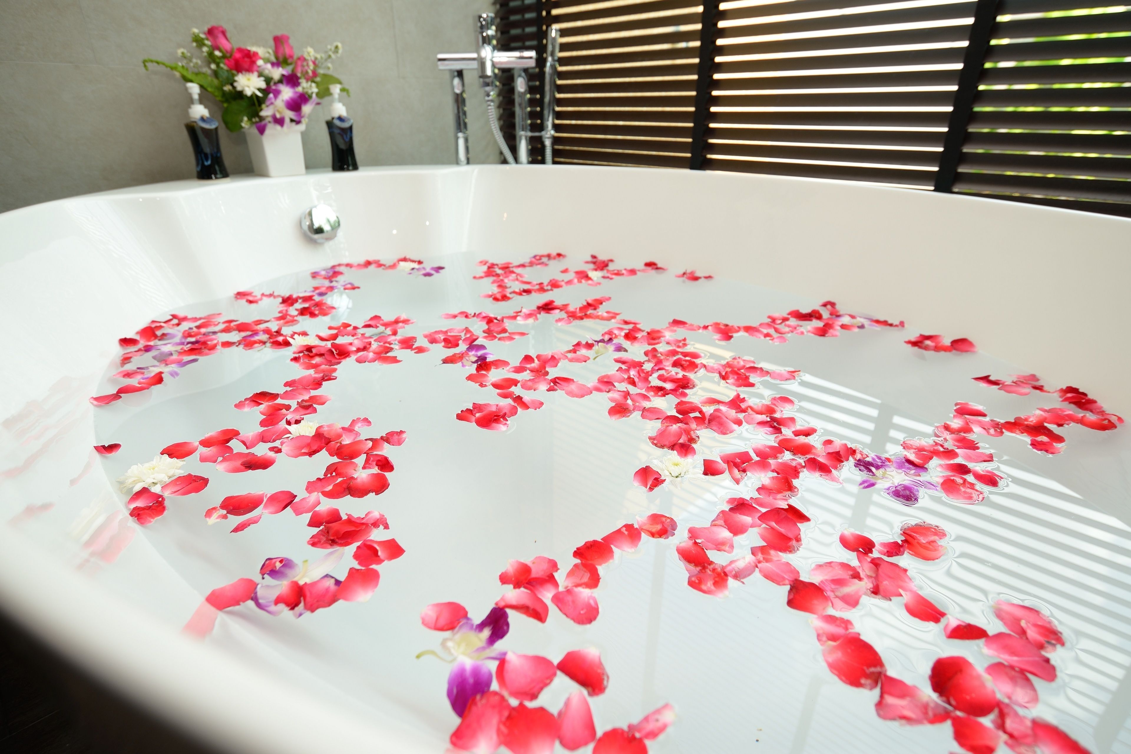 A bathtub filled with rose petals and a few flowers, with a window and blinds in the background.