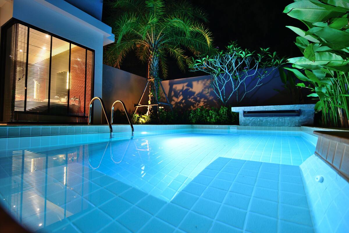A swimming pool with a tiled deck, surrounded by lush greenery and a modern building with large windows.