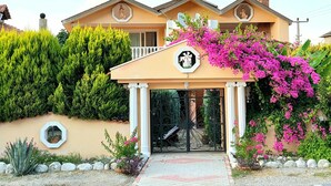 Une maison à deux étages dotée d’un porche, d’un balcon et d’un jardin où poussent différentes plantes et fleurs.