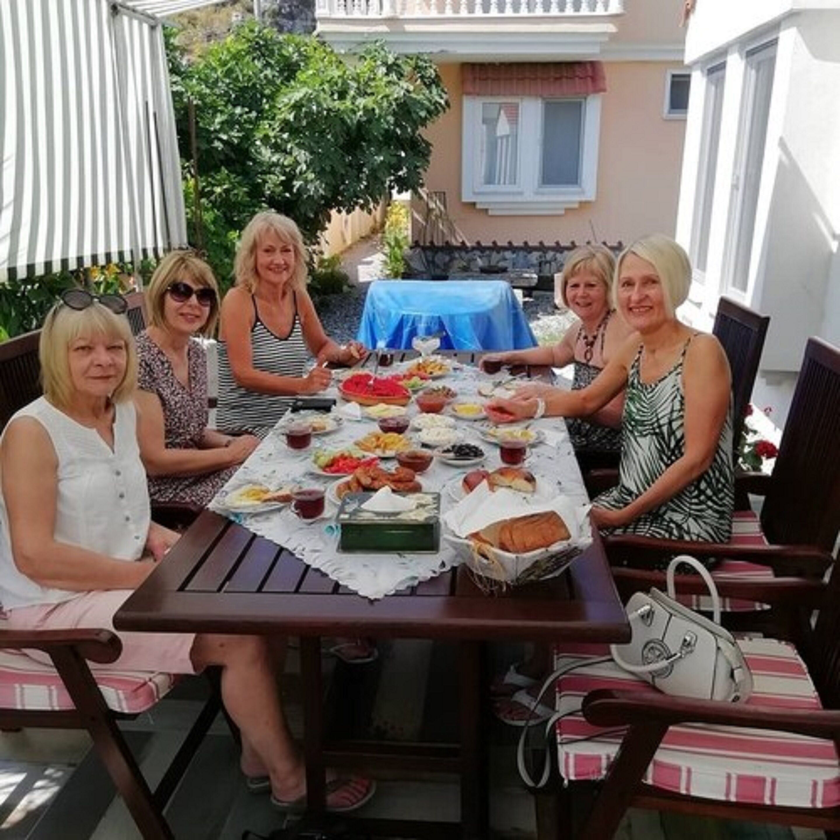 A group of people seated around a table with various dishes, in an outdoor setting with a blue tablecloth and chairs.