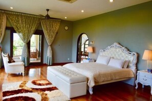A spacious bedroom with a large bed, a sitting area with a chair, and a view through a doorway.
