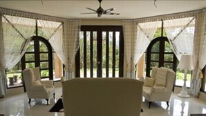 A room with a large bed, two armchairs, and a floor lamp. There are large windows with curtains and a view of greenery outside.