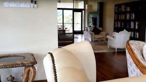 A spacious living room with a large window, a white sofa, a white armchair, a wooden coffee table, and a bookshelf filled with books.