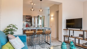 Una sala moderna con un sofá azul, un comedor con taburetes de bar, una cocina con refrigerador y un televisor en un estante de madera.