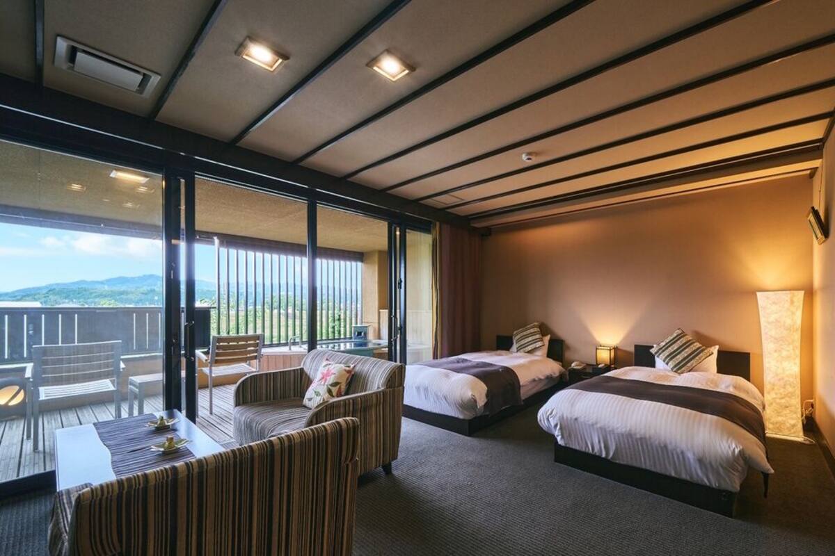 Quarto de hotel com duas camas, varanda com vista e área de refeições com mesa e cadeiras.