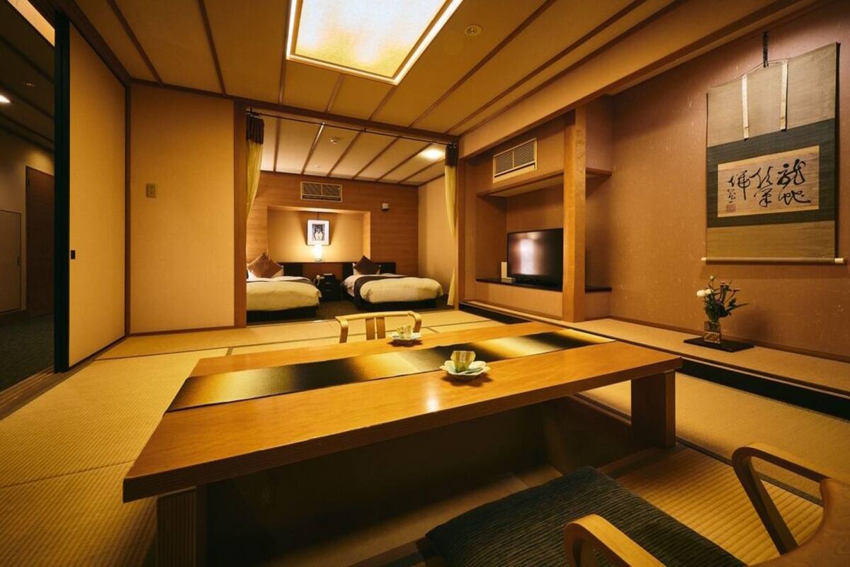 Um quarto tradicional japonês, com piso de tatame, uma mesa baixa, uma cama com futons e uma televisão.
