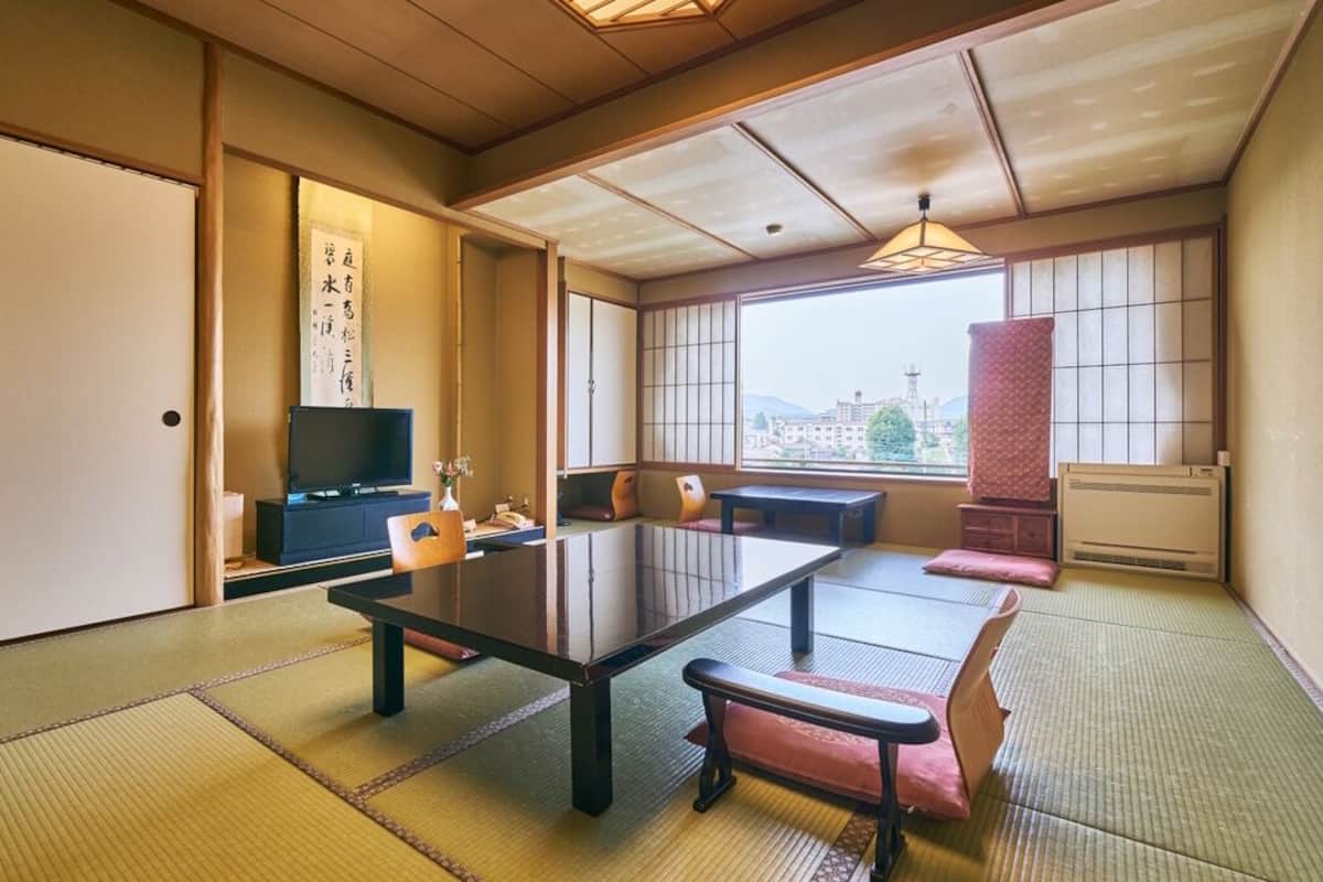 A traditional Japanese room with tatami flooring, a low table, cushions, a TV, and a view of the city.