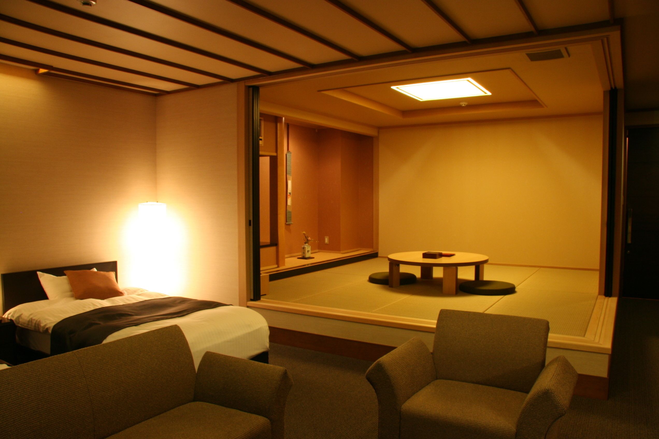 Une chambre d’hôtel comprenant un lit, deux fauteuils, une petite table et une vue sur une pièce de style japonais traditionnel, visible à travers une porte coulissante.